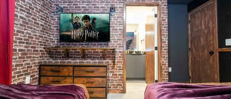 A room with a brick wall, a flat-screen TV displaying 'Harry Potter', a wooden dresser, and two beds with purple blankets.