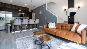 A modern living room with a brown leather sofa, two wooden coffee tables, and a dining area with bar stools.