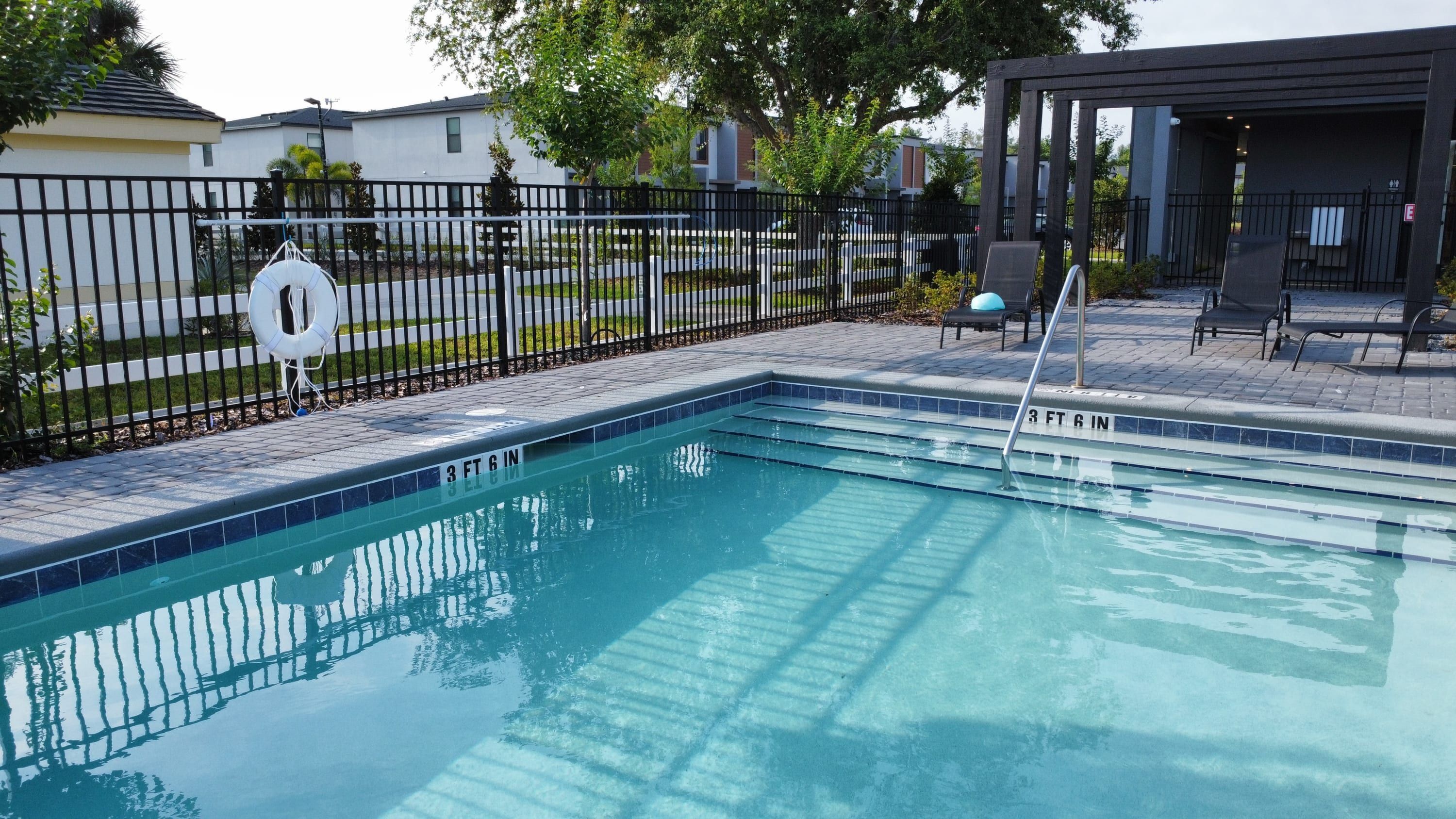 A swimming pool with a 3 ft 6 in depth marker, surrounded by a black fence, with lounge chairs and a covered seating area in the background.