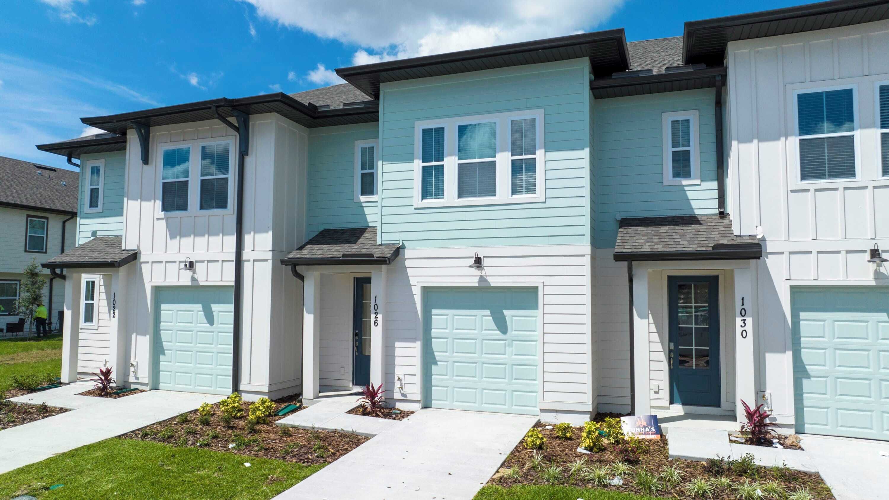 A row of townhouses with light blue siding, white trim, and dark blue garage doors. Each house has a front yard with small plants and a sidewalk.