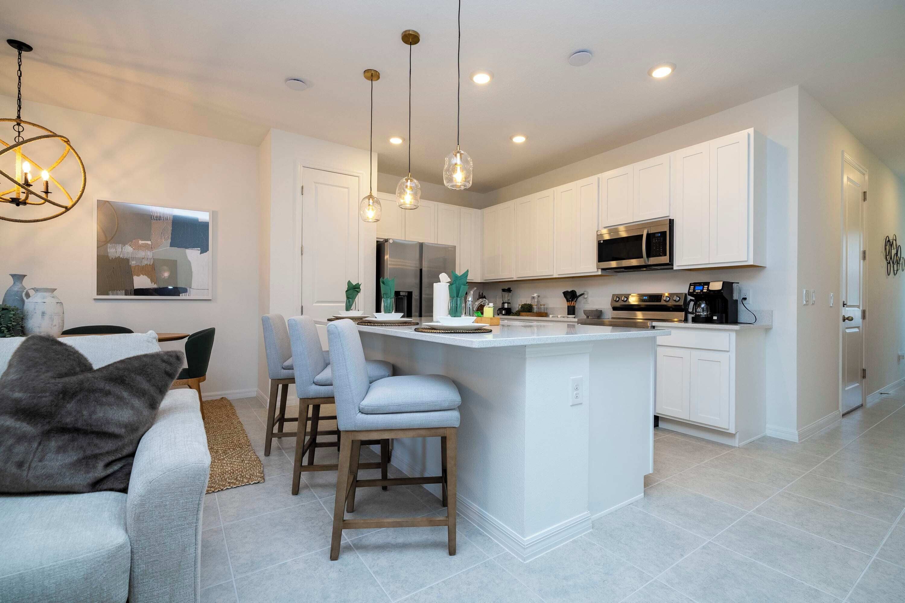 A modern kitchen with white cabinets, a central island, pendant lighting, and a dining area with a sofa and chairs.