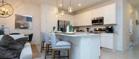 A modern kitchen with white cabinets, a central island, pendant lighting, and a dining area with a sofa and chairs.