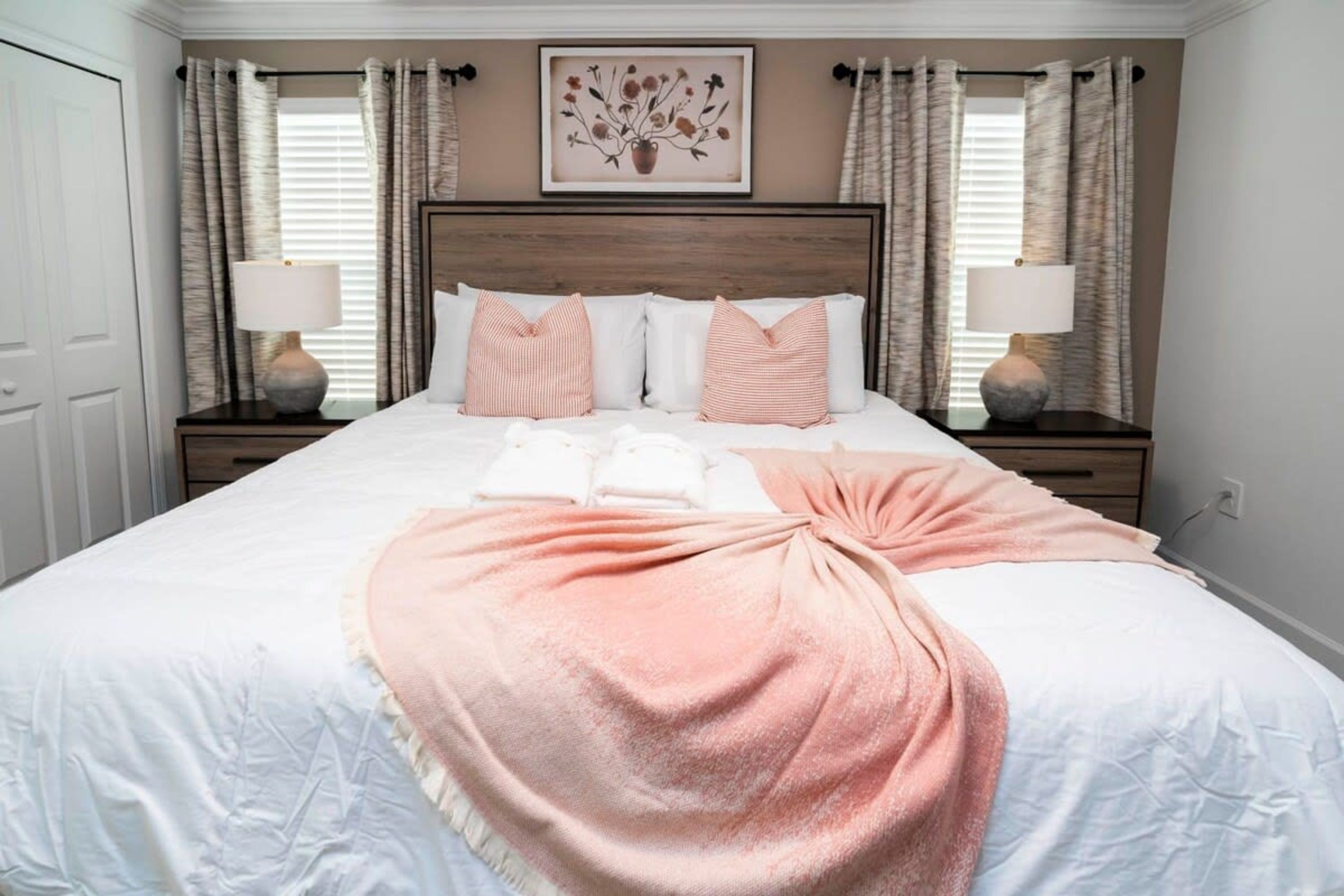 A neatly made bed with a pink throw blanket, white pillows, and a framed floral artwork above it.