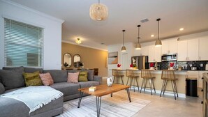 A modern living room with a grey sofa, a wooden coffee table, and a kitchen area with white cabinets and bar stools. - Wonderfull 4Bd With Pool at Champions Gate 1174 (Davenport)