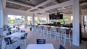 An outdoor bar area with white tables and chairs, a TV mounted on the wall, and a pool visible in the background. - Wonderfull 4Bd With Pool at Champions Gate 1174 (Davenport)