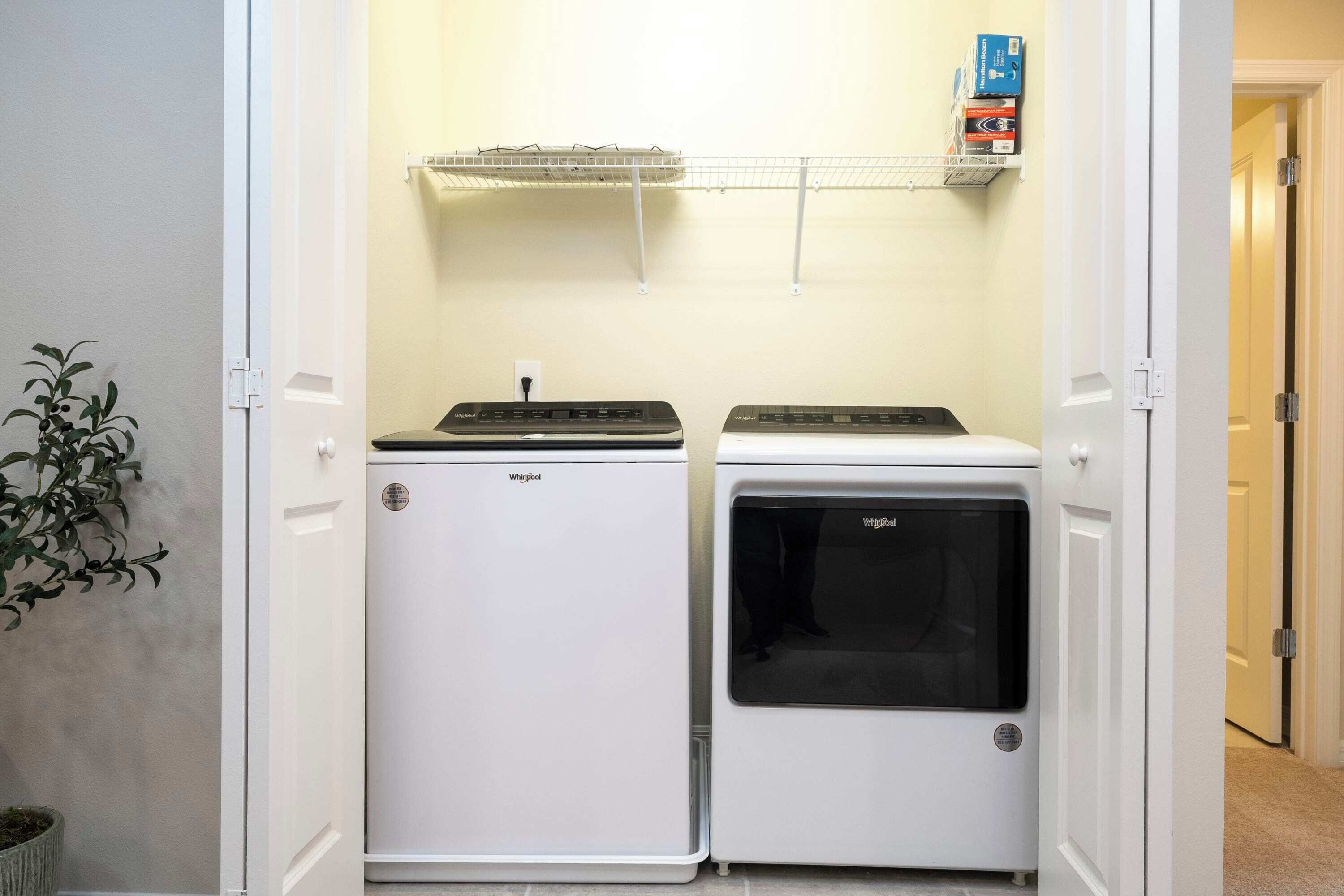 A laundry room with two stacked washers and dryers, a shelf above, and a potted plant to the left.