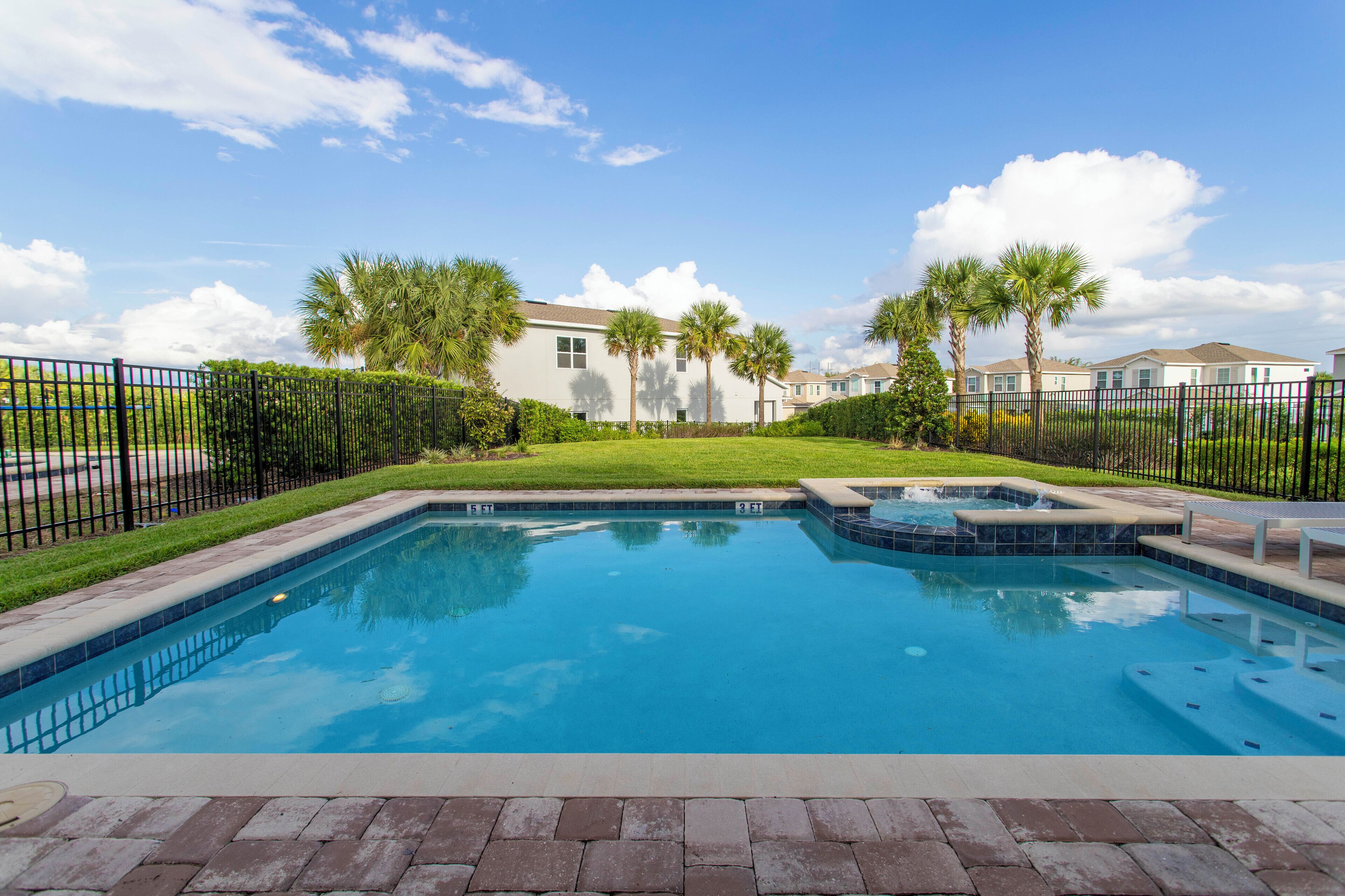 A rectangular swimming pool with a hot tub, surrounded by a brick deck, palm trees, and a fence.