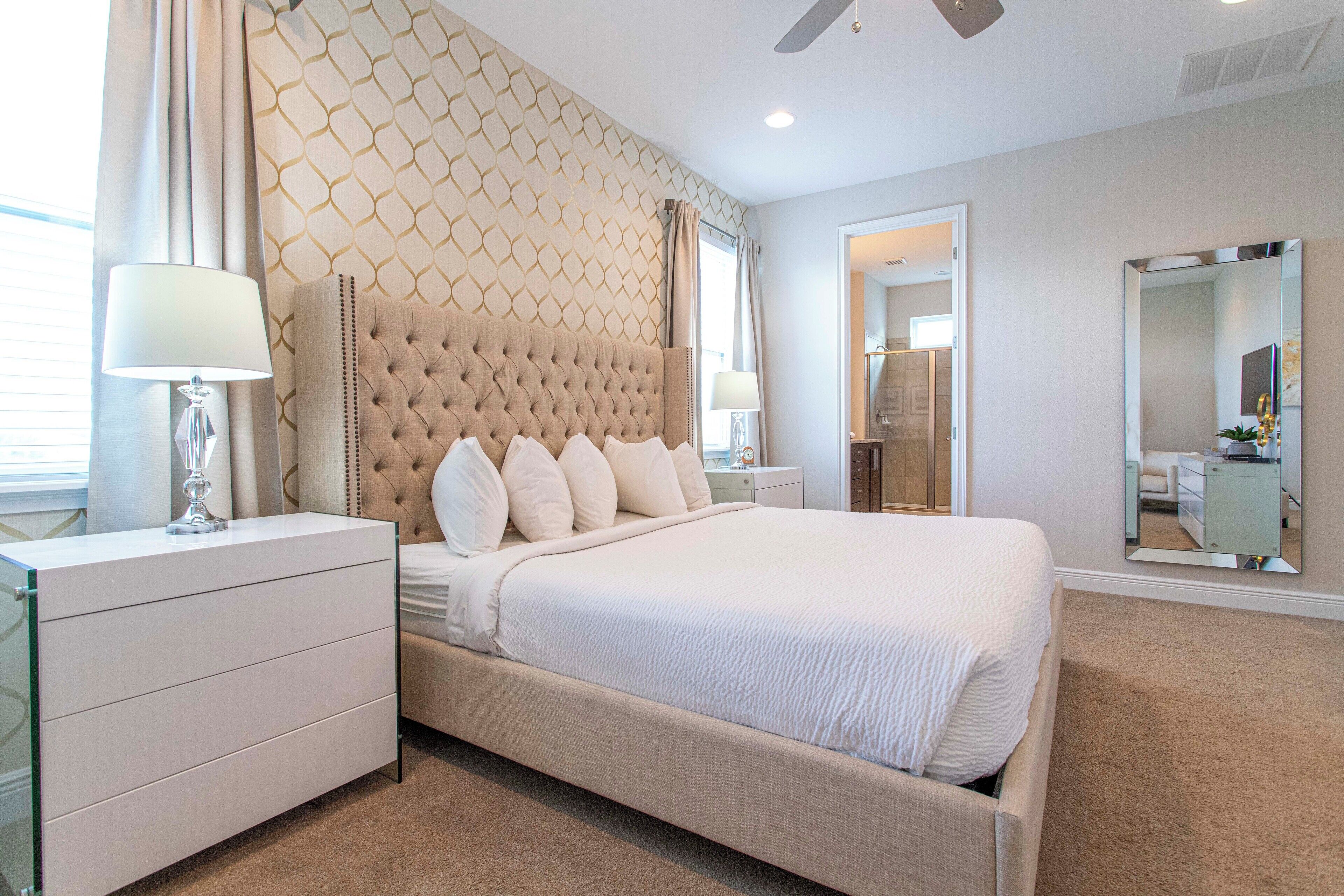 A bedroom with a tufted headboard, a large bed, a nightstand with a lamp, a dresser, and a mirror.