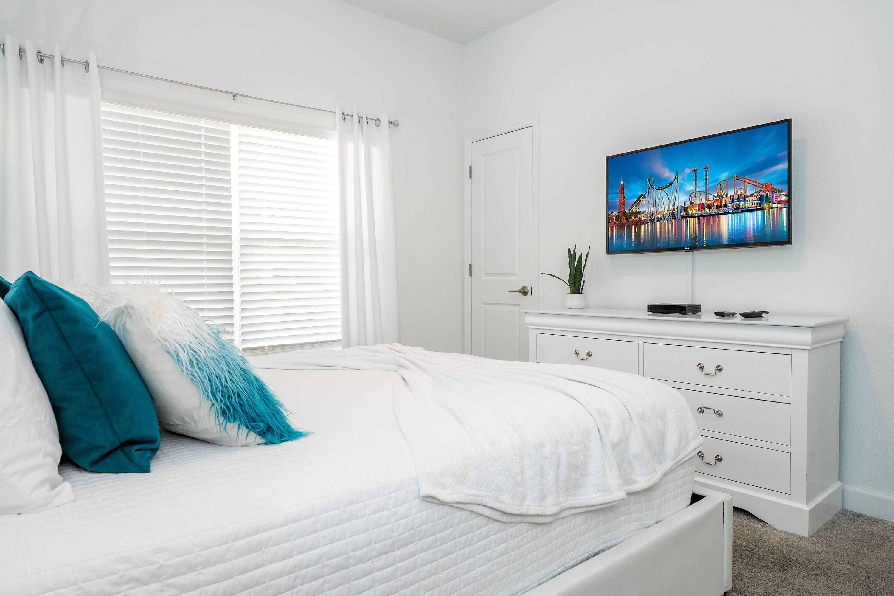 Un dormitorio con una cama, cortinas blancas, un televisor en la pared y un aparador blanco.
