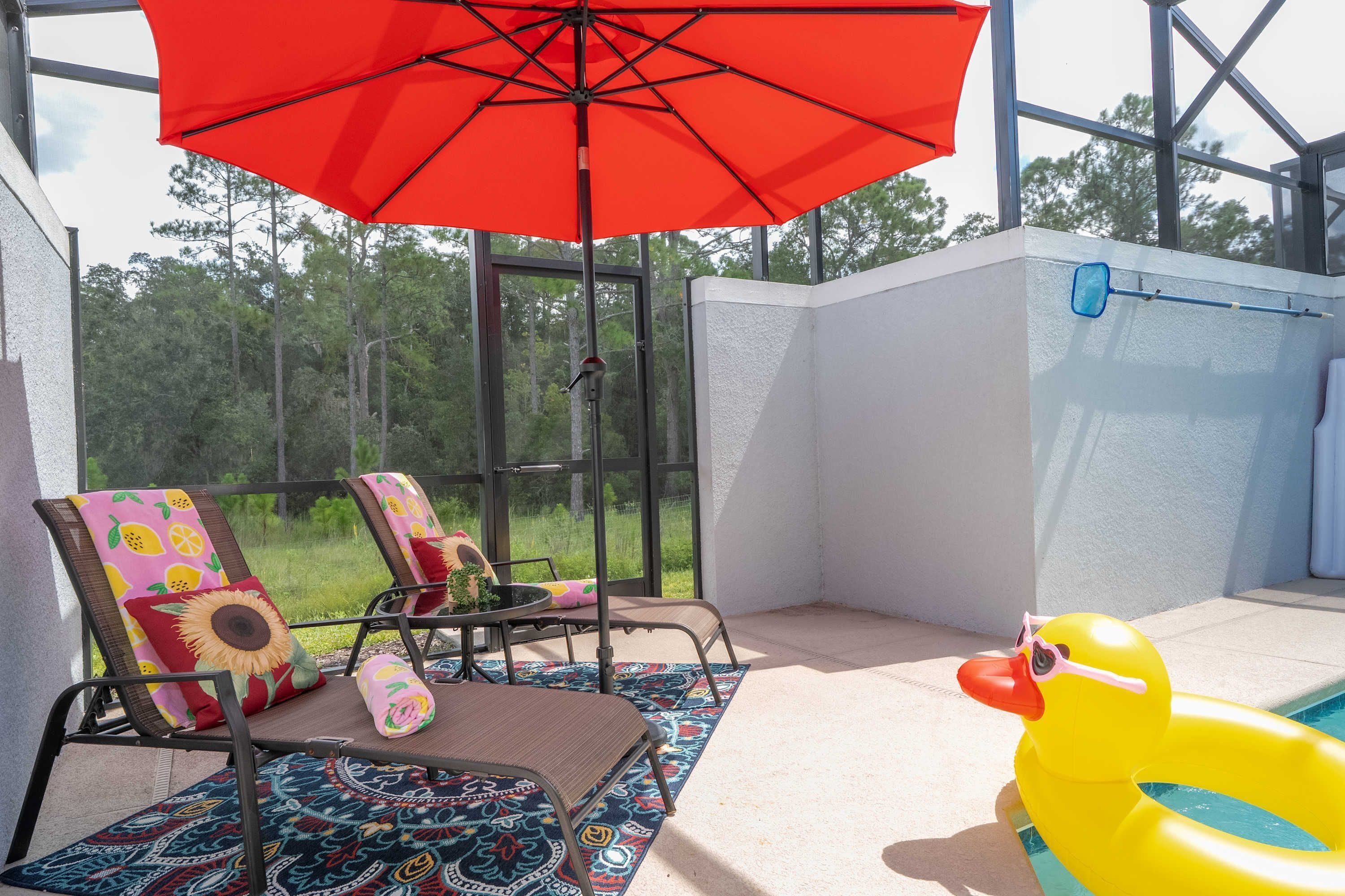 A sunlit patio with a red umbrella, two lounge chairs with colorful cushions, a pool with a yellow duck float, and a view of the surrounding greenery.