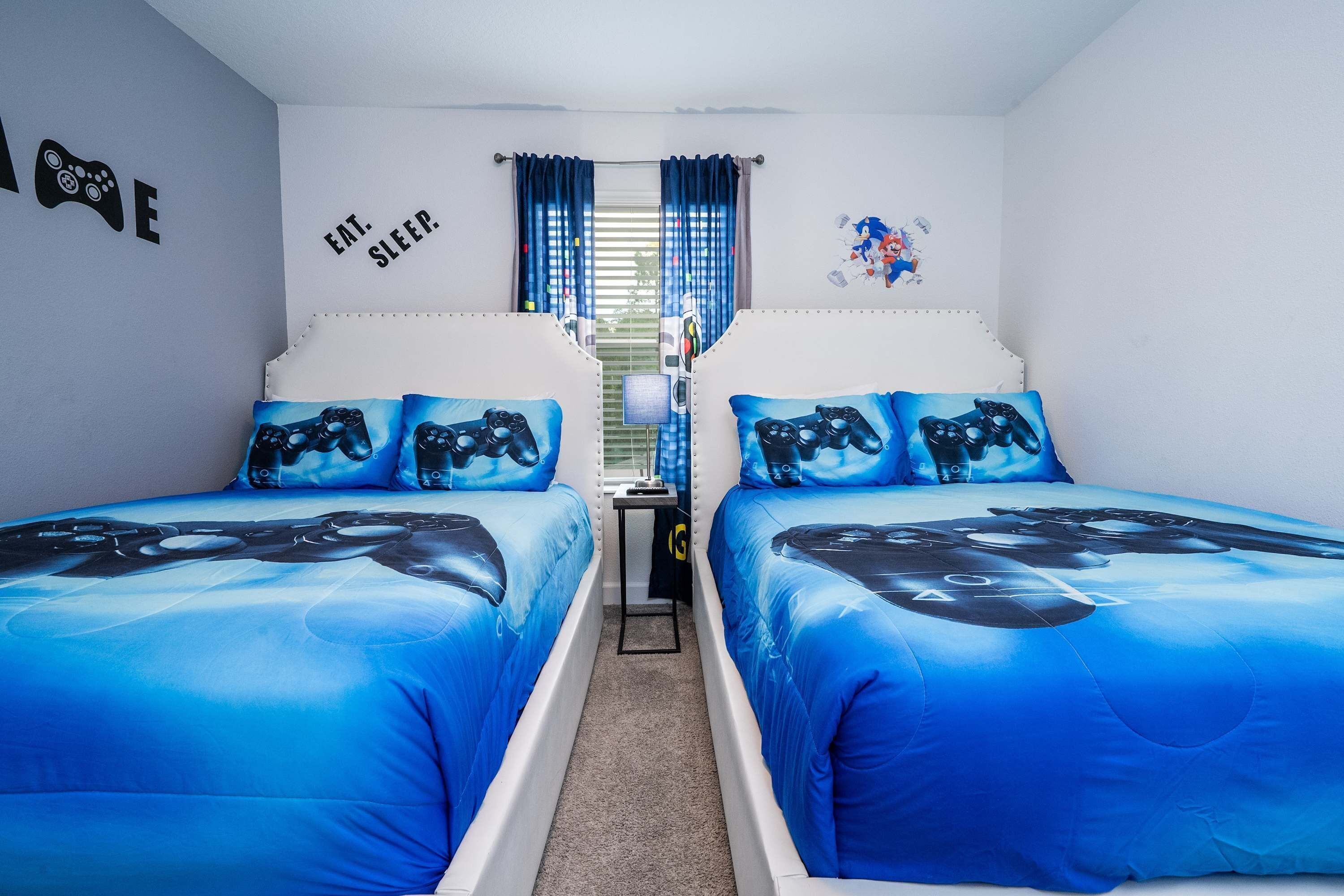 A bedroom with two beds, each with blue bedding featuring a game controller design. There is a wall with a 'Eat. Sleep.' sign and a small table with a lamp.