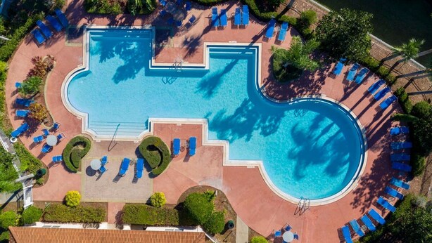 Vista aérea de um resort com uma piscina grande central, várias espreguiçadeiras e vegetação ao redor.