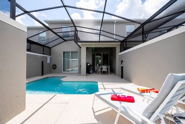 A swimming pool with lounge chairs, a glass-enclosed patio, and a view of a building in the background.