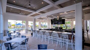 A spacious outdoor dining area with white tables and chairs, a bar with stools, and a pool in the background.