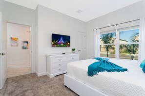 A bedroom with a large bed, a white dresser, a TV on the wall, and a window with a view of palm trees.