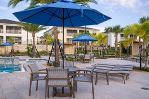 A poolside area with lounge chairs and umbrellas, surrounded by palm trees and a building in the background.