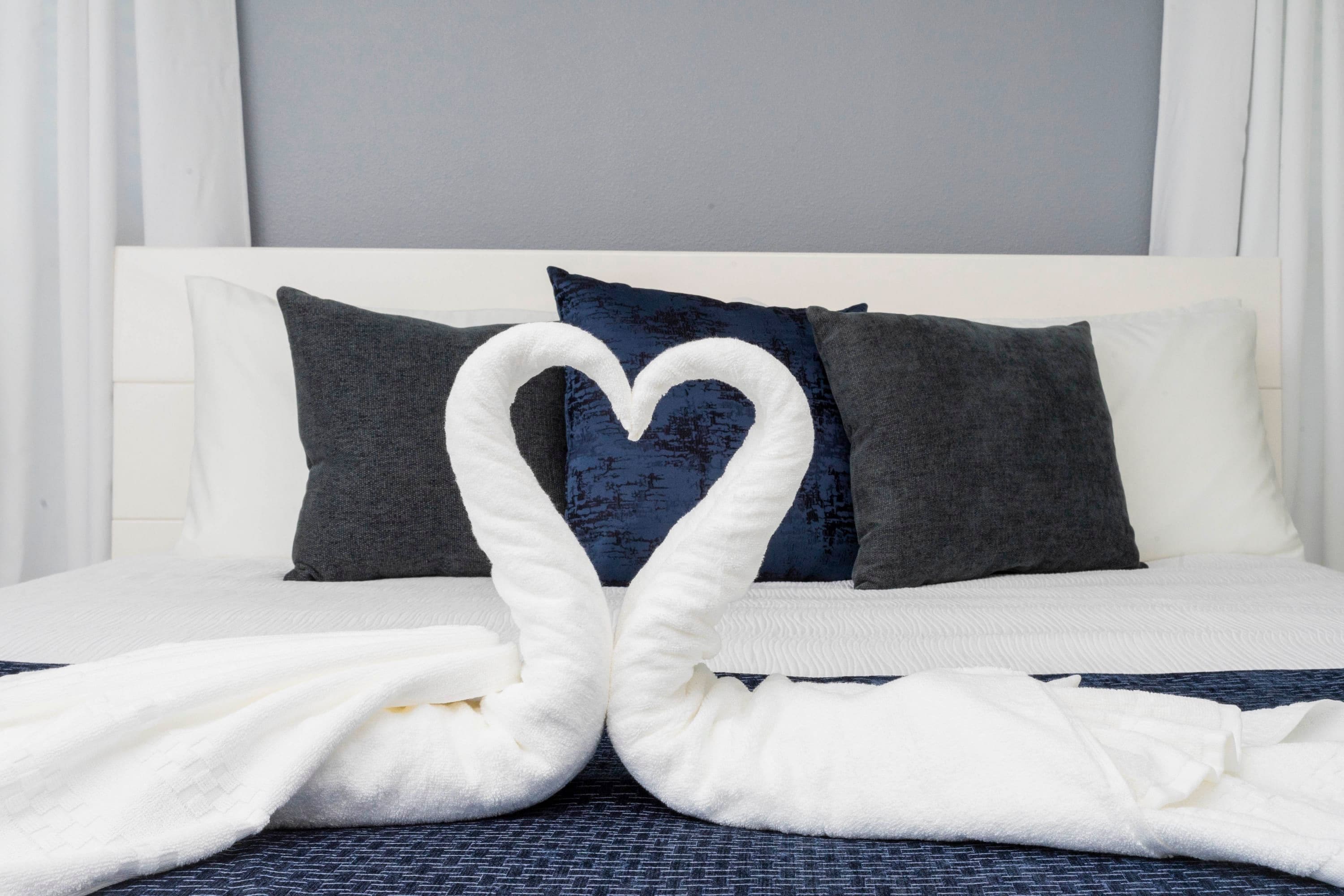 A neatly made bed with a heart-shaped towel arrangement in the center, white bedding, and dark blue pillows.
