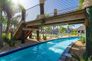 Un espace piscine agrémenté d’un pont en bois, de palmiers et d’un ciel dégagé.
