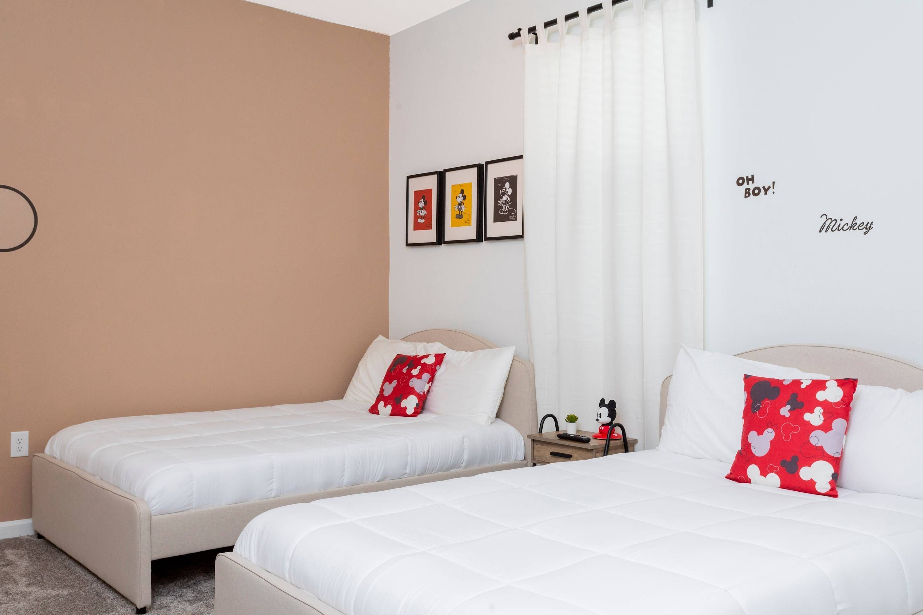 Quarto de hotel com duas camas, uma pequena mesa de cabeceira com uma planta, parede com quadros e uma cortina.