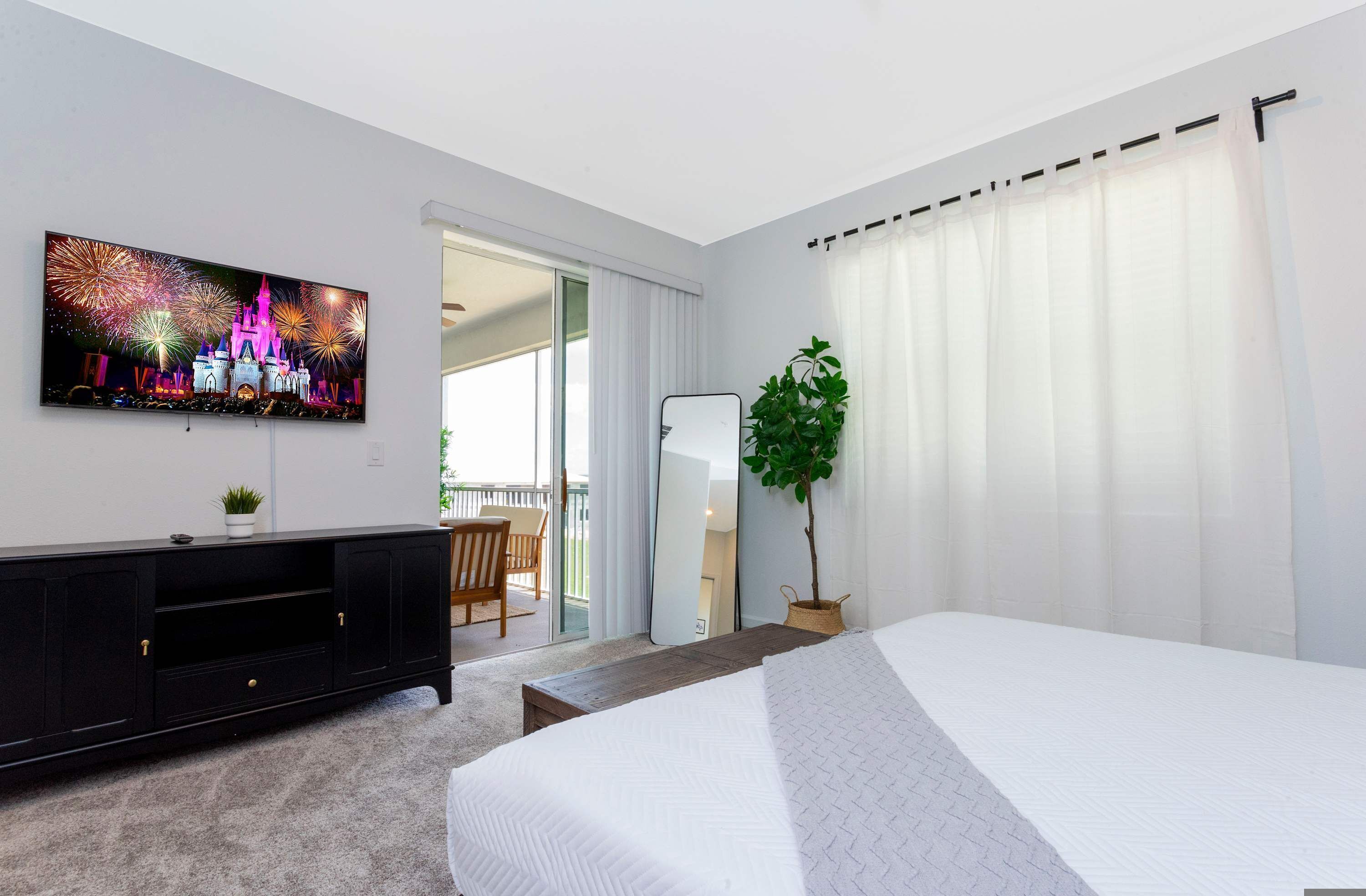 A hotel room with a bed, a TV mounted on the wall, a plant, and a balcony with a view.