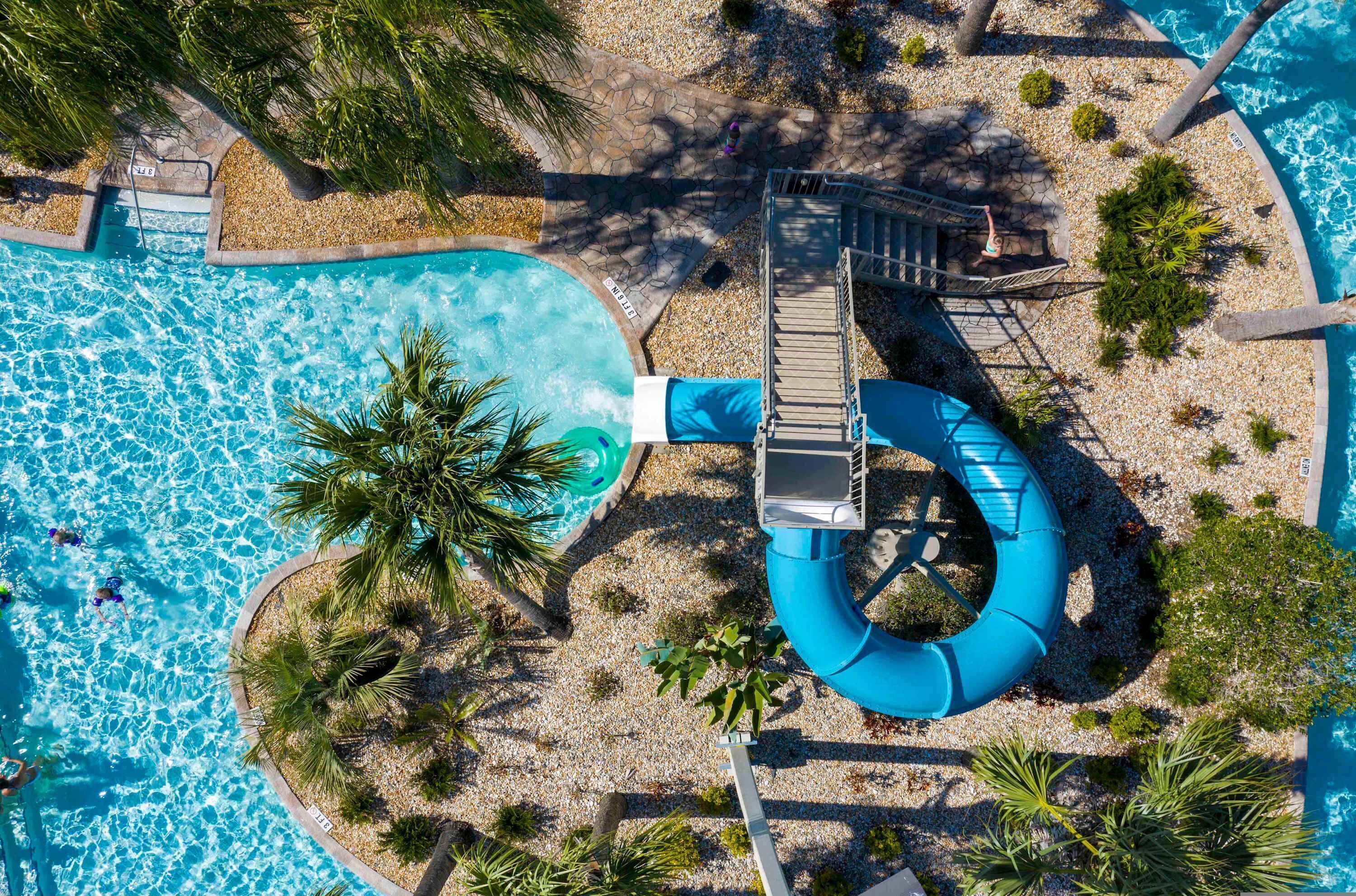 Una vista aerea di una piscina con uno scivolo, circondata da palme e una spiaggia di ciottoli.