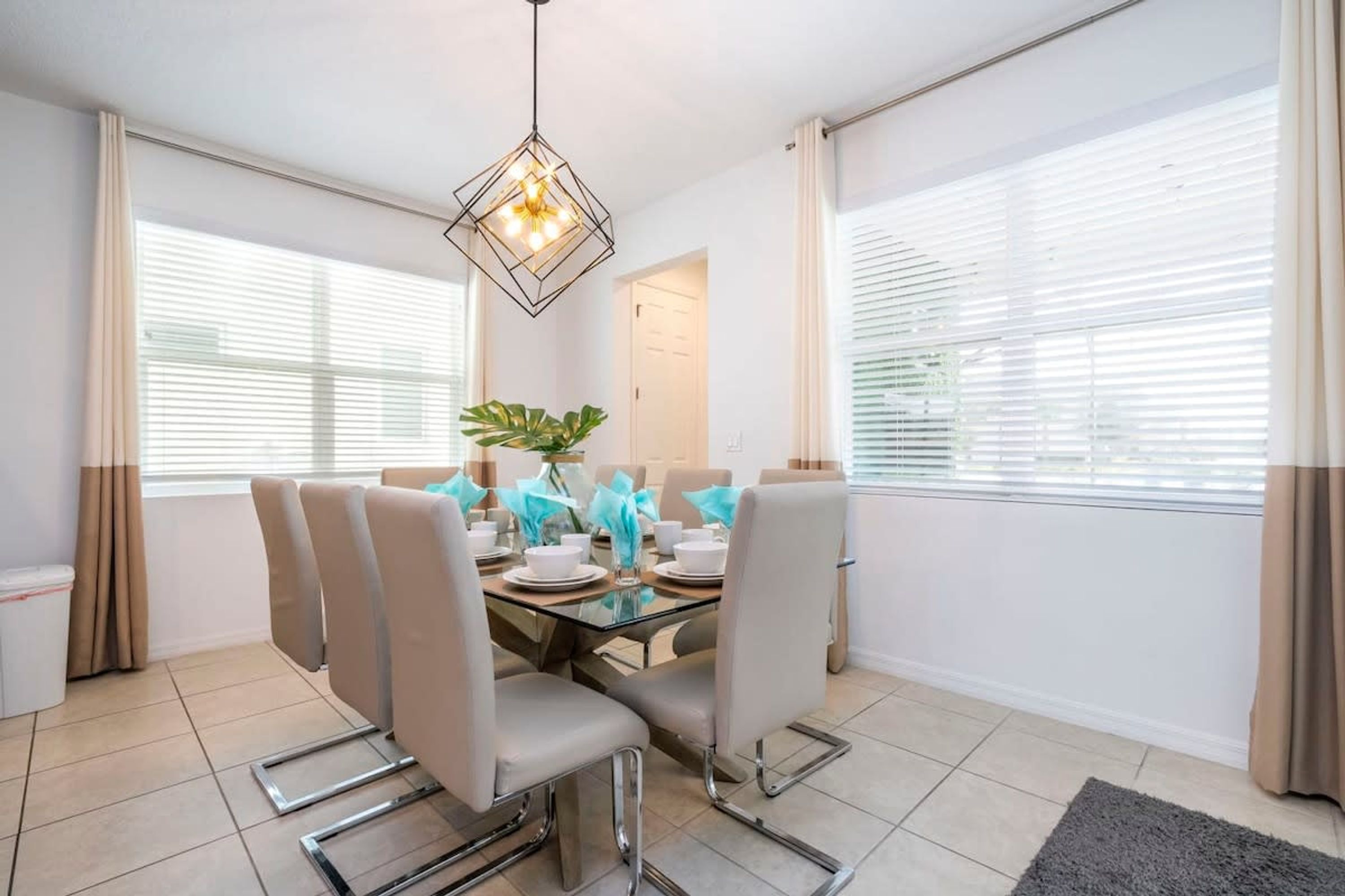 A dining area with a glass table, six chairs, a hanging light fixture, and large windows with blinds.
