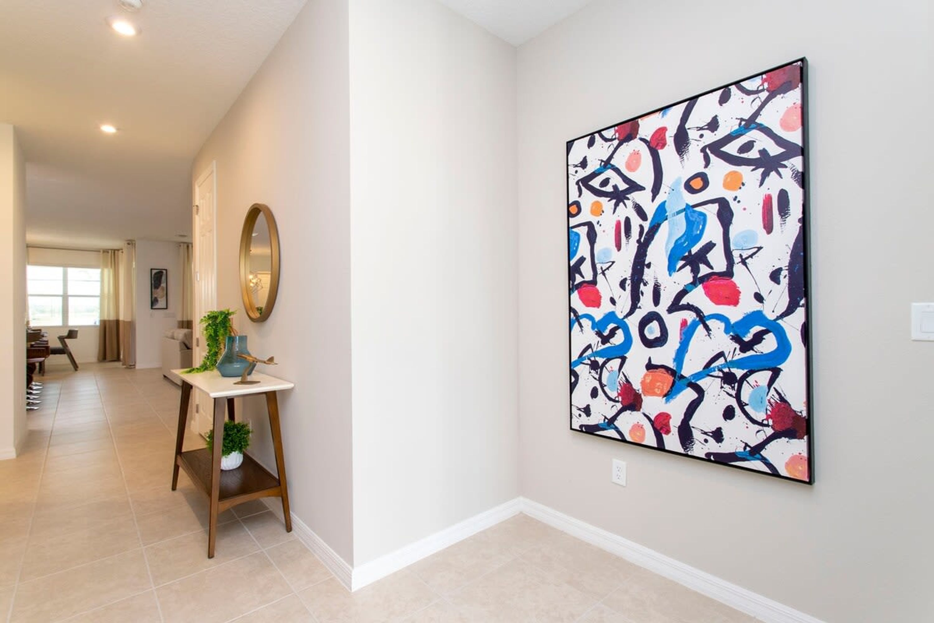 A hallway with a large abstract painting on the wall, a round mirror, and a small table with a vase and plant.