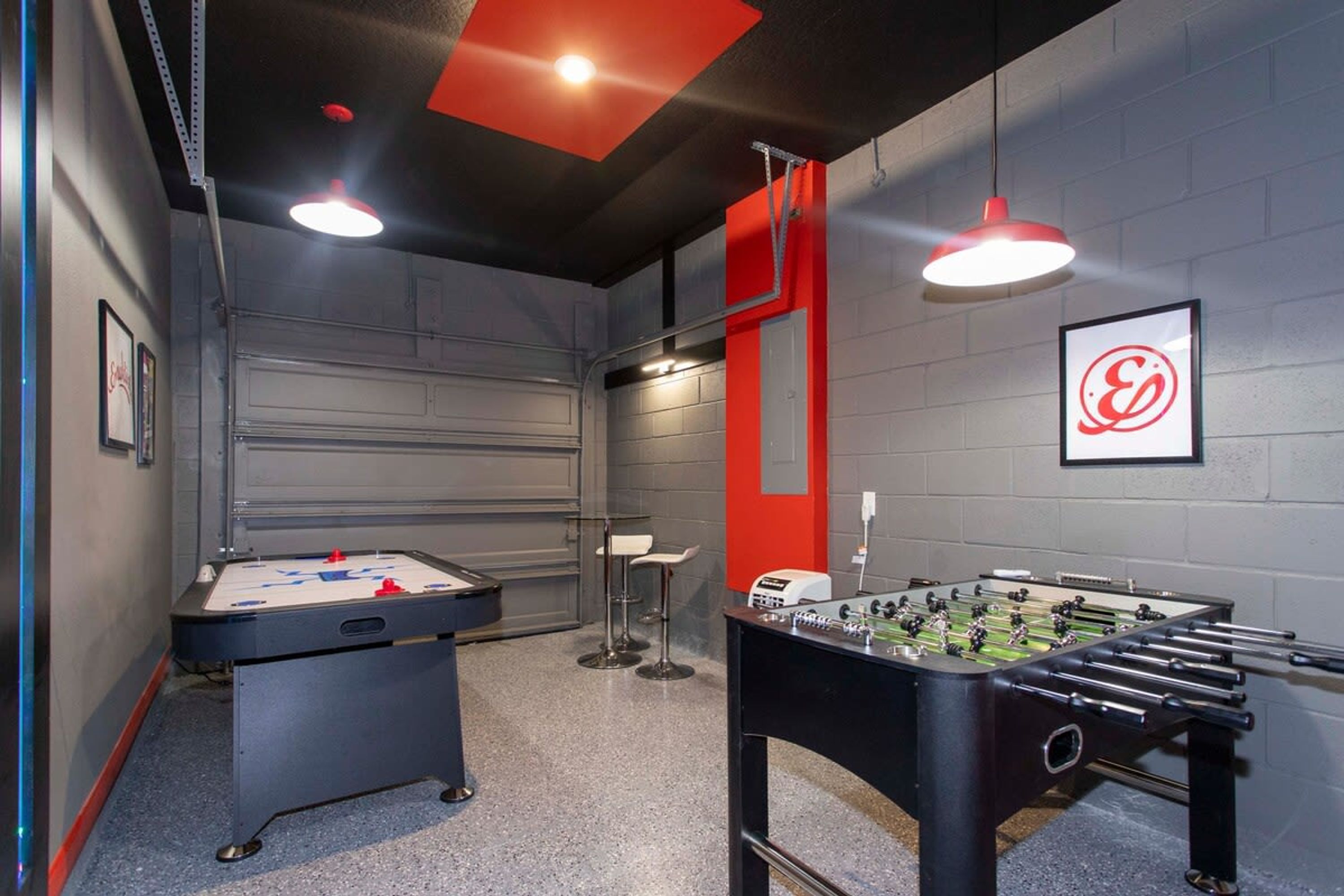 Una habitación con dos mesas de fútbolín, una zona de bar con taburetes y una pared de acento roja con un logotipo.