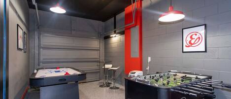 Una habitación con dos mesas de fútbolín, una zona de bar con taburetes y una pared de acento roja con un logotipo.
