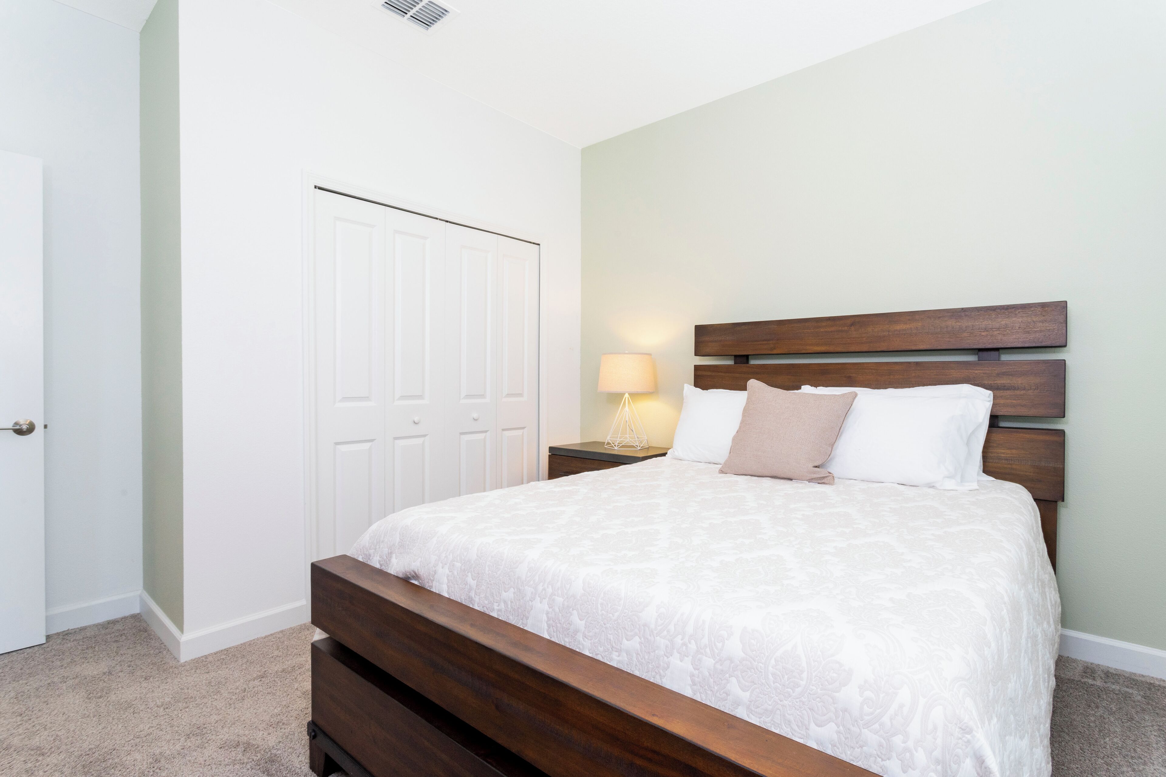 A bedroom with a wooden bed, white bedding, a nightstand with a lamp, and a white door.