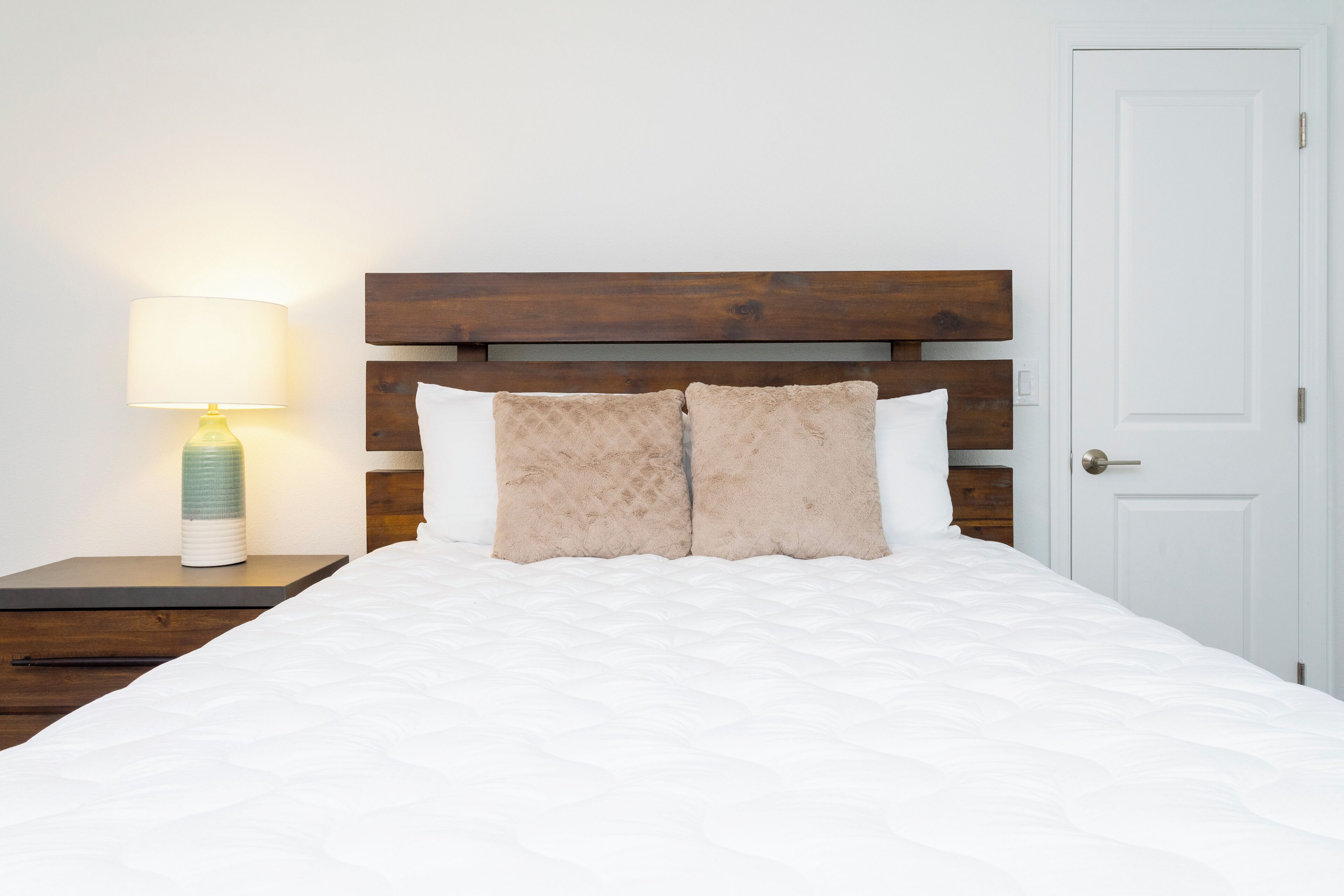 Una cama bien tendida con un cabecero de madera, una lámpara de noche y una puerta blanca al fondo.