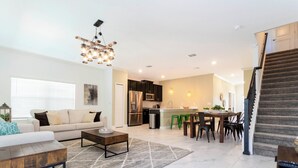 Ein geräumiger Wohnbereich mit einer Couch, einem Couchtisch und einem Essbereich mit Stühlen und Tisch. Im Hintergrund ist eine Küche mit modernen Geräten zu sehen.