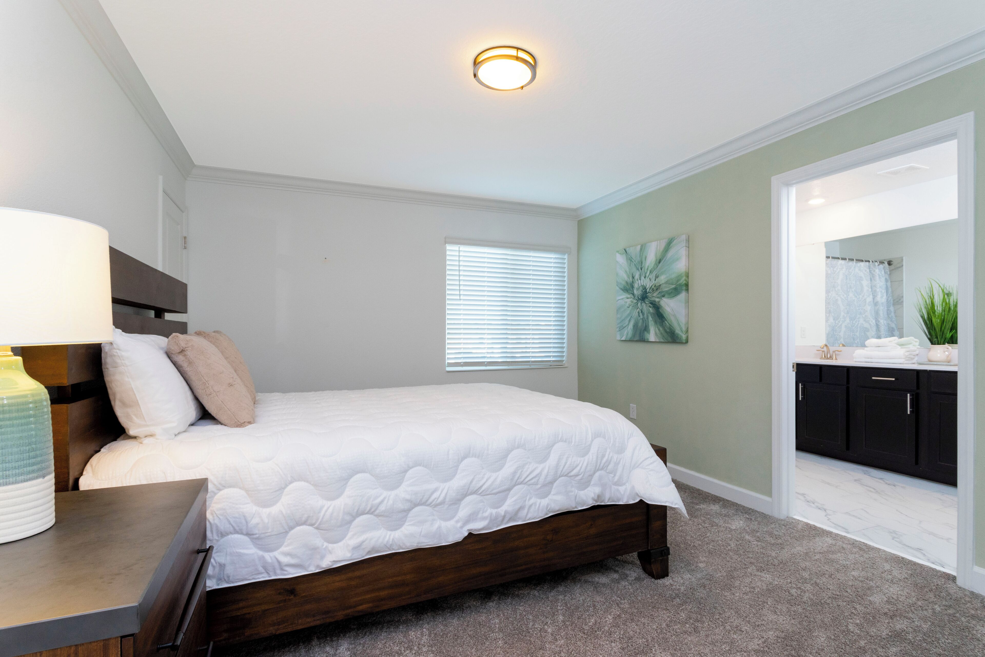 A bedroom with a bed, bedside table, lamp, window, and a view into the bathroom.