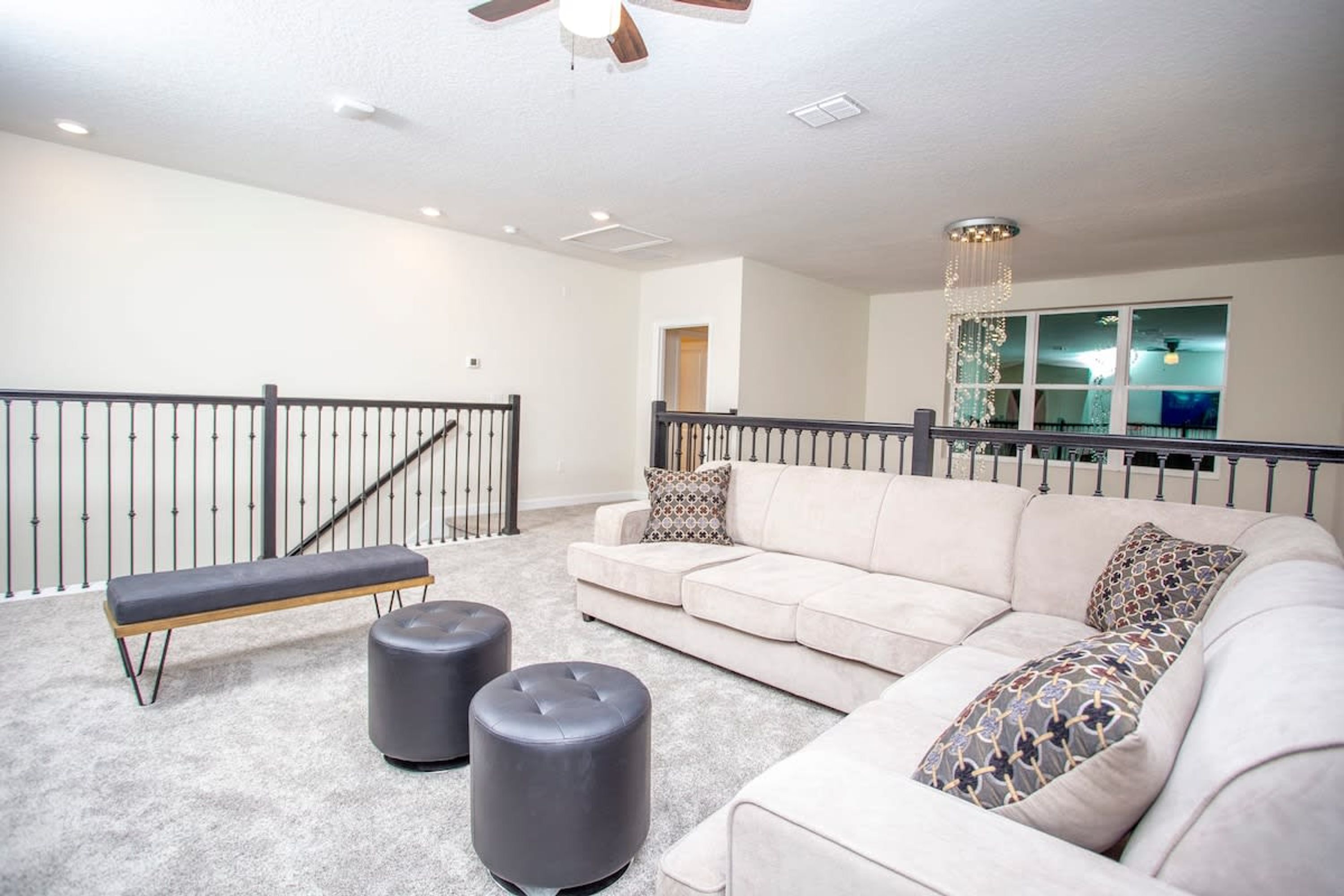 A spacious living room with a beige sofa, two ottomans, a bench, and a staircase with a railing.