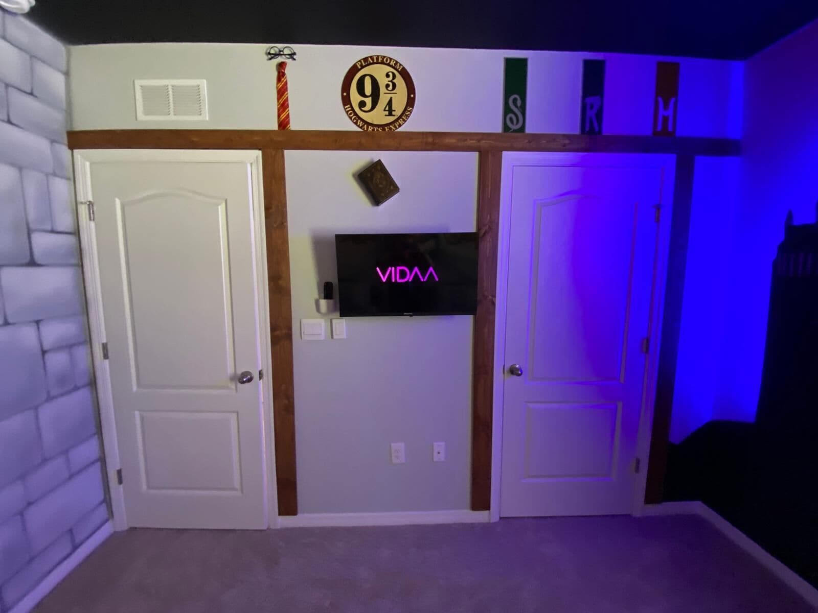 A room with a white brick wall, a door, a TV mounted on the wall, and a sign above the door with the number 9.