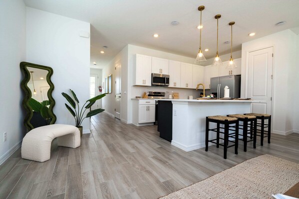 A modern kitchen with white cabinets, a black refrigerator, and a microwave. There are three bar stools and a large plant.