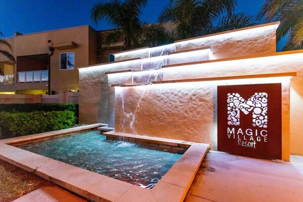 Un élément décoratif aquatique avec de l’eau en cascade, une piscine et un panneau indiquant « MAGIC VILLAGE RESORT ».