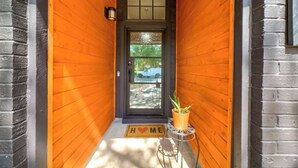 Un couloir extérieur en bois, avec une porte noire, une petite plante en pot et un paillasson sur lequel est écrit « ENTRÉE ».