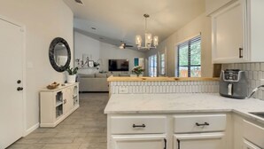 Une cuisine moderne avec des meubles blancs, un plan de travail en marbre et un îlot central. On y trouve un ventilateur de plafond, un lustre et une vue sur un salon avec un canapé et un téléviseur.
