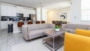 Une cuisine moderne avec des meubles blancs, un four à micro-ondes et un réfrigérateur. On y trouve également un coin repas avec un canapé gris et un fauteuil jaune.