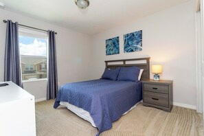 Um quarto com cama, mesa de cabeceira com luminária, escrivaninha e janela com cortinas.