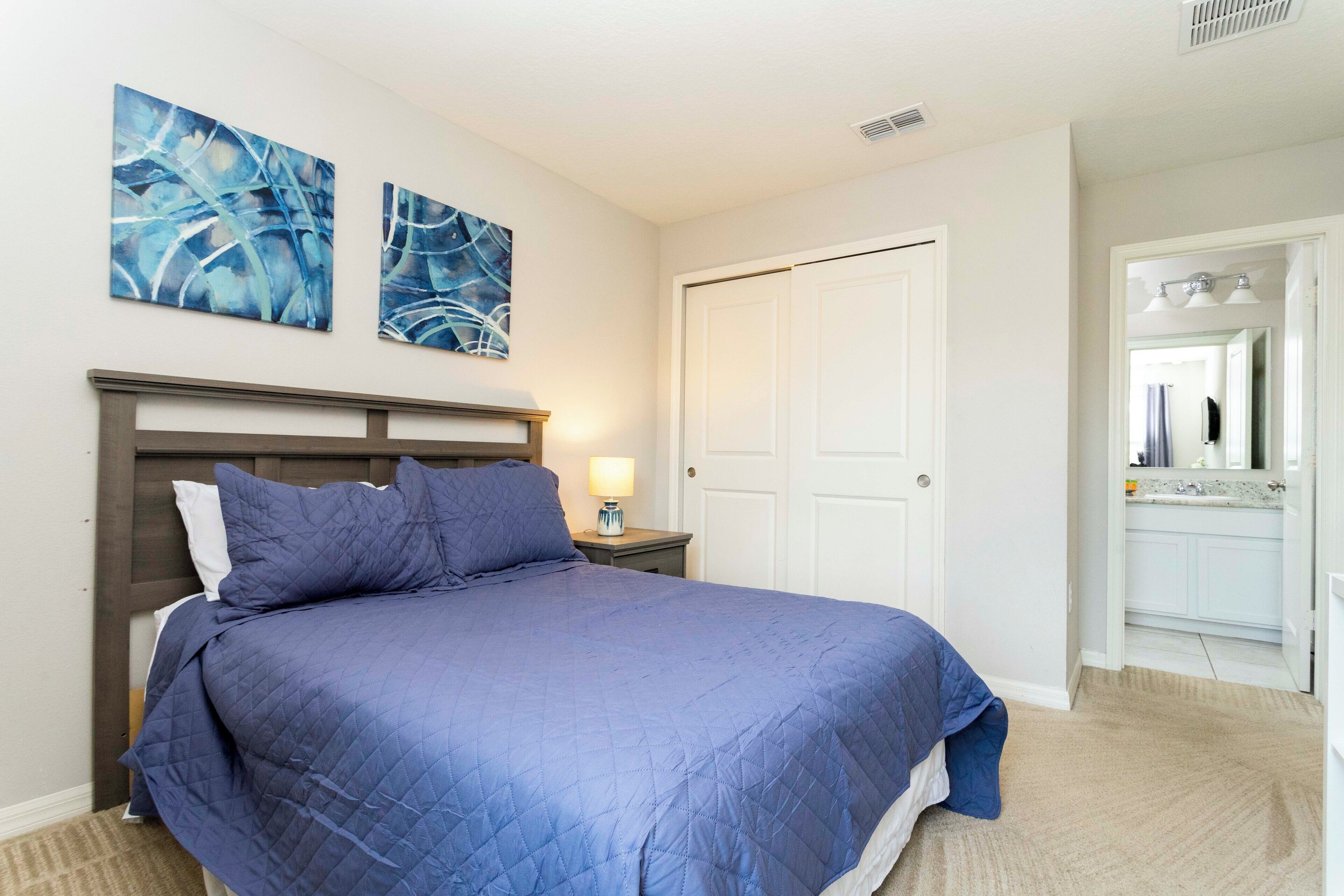 A bedroom with a bed, two abstract paintings, a nightstand, a lamp, a door, and a bathroom visible through an open door.
