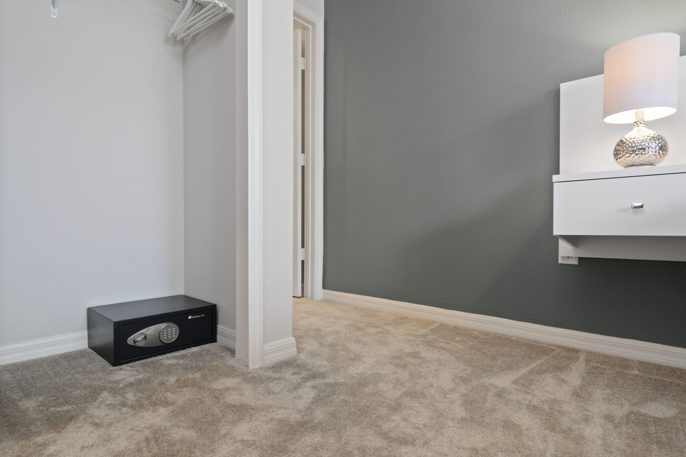 Ein Zimmer mit grauer Wand, einem weißen Nachttisch mit Lampe und einem schwarzen Safe.