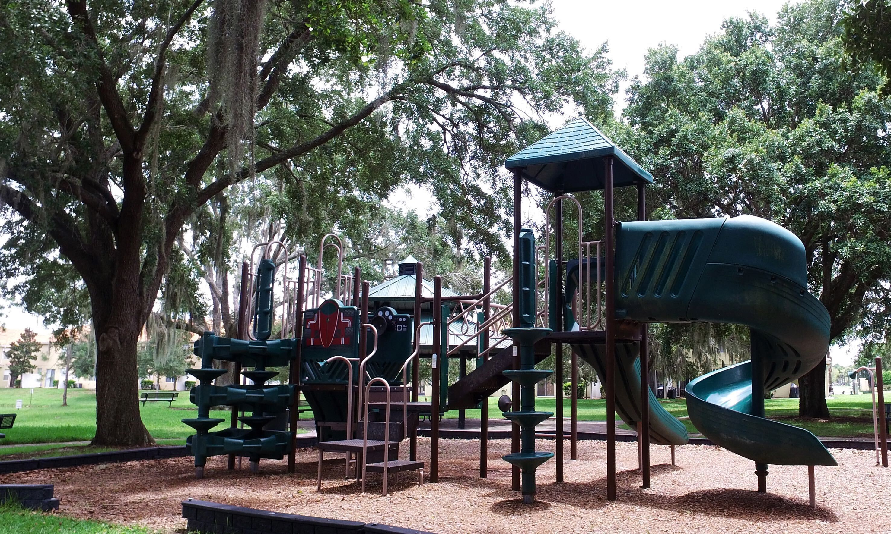 A playground with a slide and climbing structures, surrounded by trees and a grassy area.