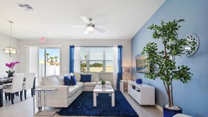 Una sala moderna con un sofá blanco, una mesa de centro y un tapete azul. Se observa un comedor con una mesa y sillas, y una planta en una maceta azul.