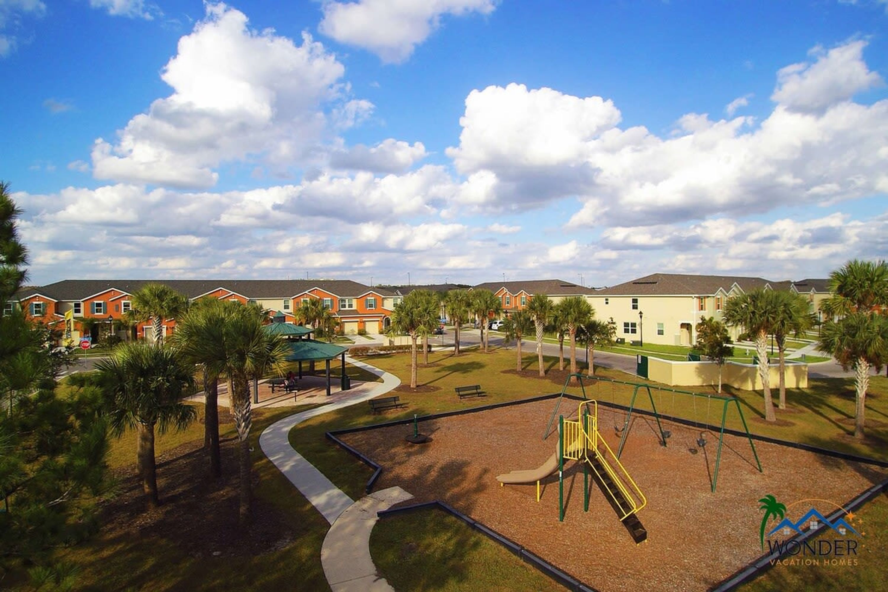 A playground with swings and a slide, surrounded by palm trees and a walking path.