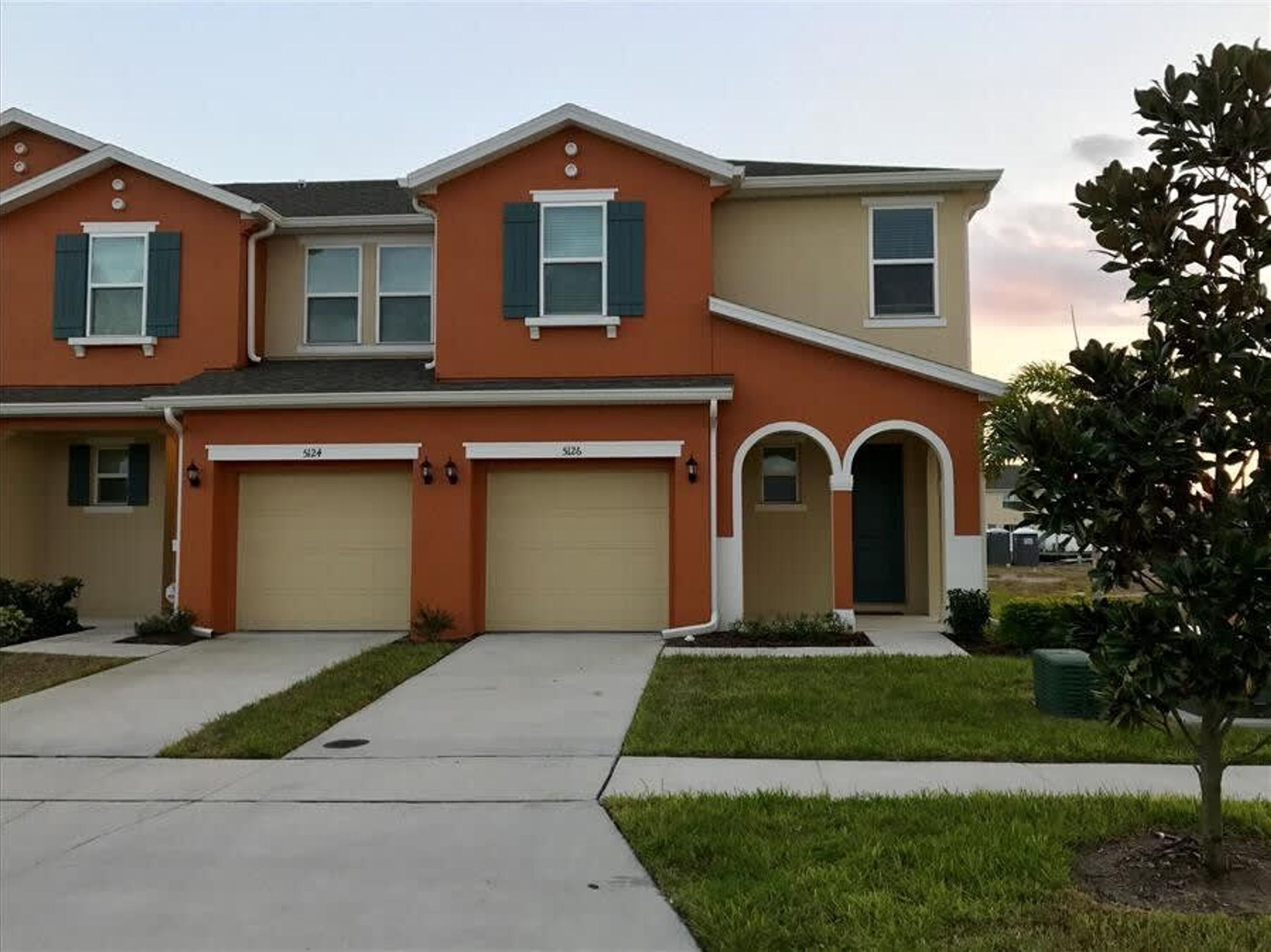 A two-story residential building with a beige and orange facade, two garage doors, and a small front yard with a tree.