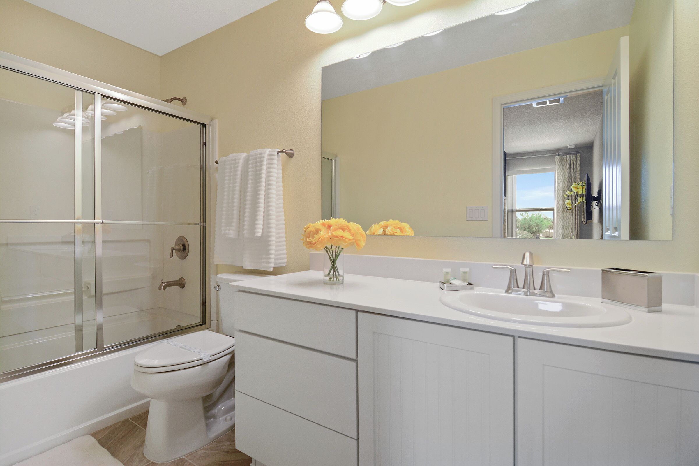 A modern bathroom with a glass-enclosed shower, a white toilet, a large mirror, and a white sink with a vase of yellow flowers.