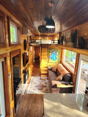 A cozy cabin interior with a loft bed, wooden walls, a dining area, and a view of the outdoors.
