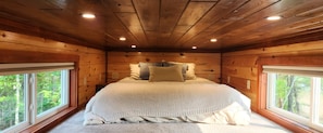 Cozy cabin-style bedroom with a large bed, wooden walls, and forest views through two windows.